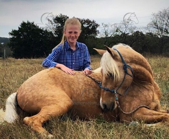Young girl smiling and sitting next to a palomino horse lying in a field.
